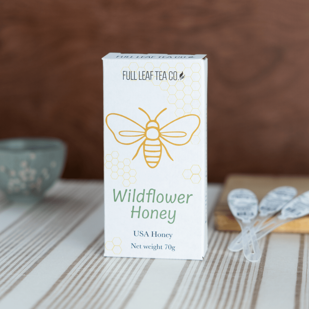 A Ten pack box of Full Leaf Tea Co. Wildflower Honey Spoons stands on a striped tabletop with Wildflower Honey Spoons and a blurred ceramic cup in the background.